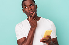 A photograph of a young man wearing a white T-shirt and holding a cell phone with a puzzled expression on a blue background