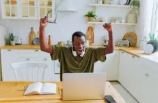 A photo of a man celebrating at a kitchen table with a laptop and headphones, surrounded by a cozy, modern kitchen.