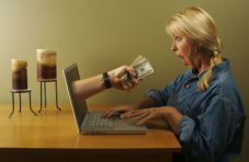 A photo of a shocked women looking at a man’s arm extending from her laptop screen holding wads of bank notes