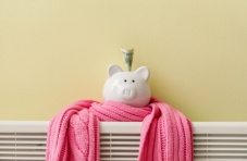 A white piggy bank with a dollar note wrapped in a pink scarf on a radiator against a light yellow wall