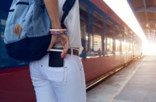 A close-up image of a woman on a subway platform with a backpack on her back taking a smartphone out of her back pocket